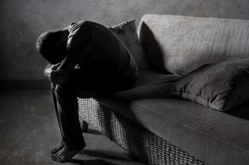 lifestyle dramatic light portrait of young sad and depressed man sitting at shady home couch in pain and depression feeling stressed and desperate