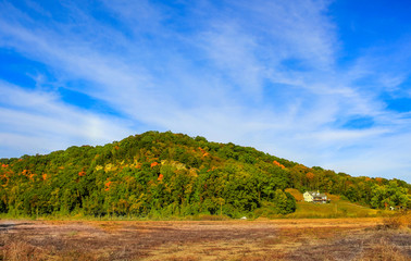 View of Midwestern bluffs covered with colorful  fall trees; dry field in the foreground; house at...
