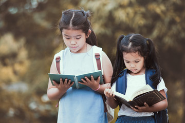 Back to school. Cute asian child girl with school bag reading a book with her sister together in nature background in vintage color tone