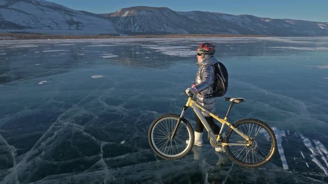 Woman is riding bicycle on the ice. The girl is dressed in a silvery down jacket, cycling backpack and helmet. The cyclist rides and stops to rest. He sits down on the wheel and partially takes off