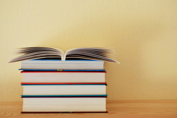 Stack of colorful books and one open book on top on wooden bookshelf in home on yellow wall...