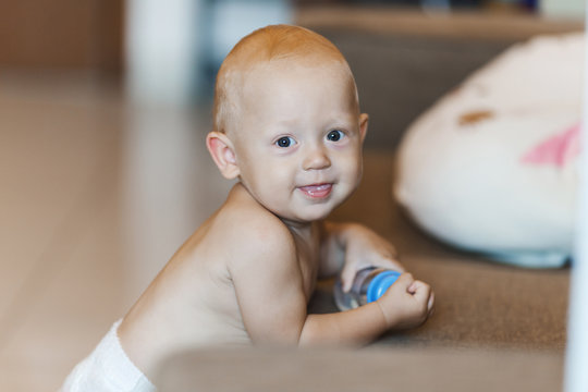 A child of 8 months is at the sofa with a bottle of water, smiling in a diaper
