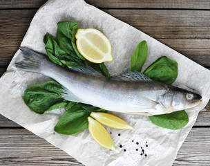 Raw pikeperch on parchment paper. Next to the fish are fresh spinach and lemon slices. Light wooden...