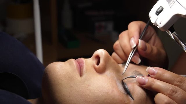 Woman Eye with Long Eyelashes. Eyelash Extension. Lashes, close up, selected focus. Gluing artificial eyelashes with tweezers. Beautician in magnifier glasses performs laborious small work.