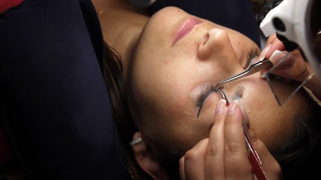 Woman Eye with Long Eyelashes. Eyelash Extension. Lashes, close up, selected focus. Gluing artificial eyelashes with tweezers. Beautician in magnifier glasses performs laborious small work.