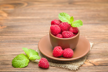 Ripe raspberries with green mint leaves in brown cup and saucer on sackcloth and wooden background.