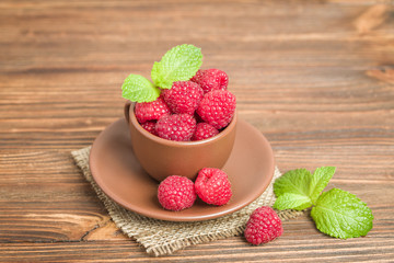 Ripe raspberries with green mint leaves in brown cup and saucer on sackcloth and wooden background.