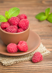 Ripe raspberries with green mint leaves in brown cup and saucer on sackcloth and wooden background.