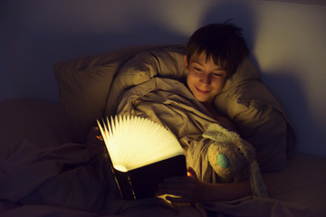 Adorable boy reads book before sleeping in bed at home. Childhood.