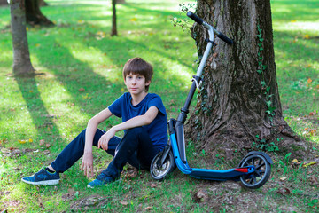 Boy sitting and resting near tree with his scooter, summer nature outdoor. image toned and noise...