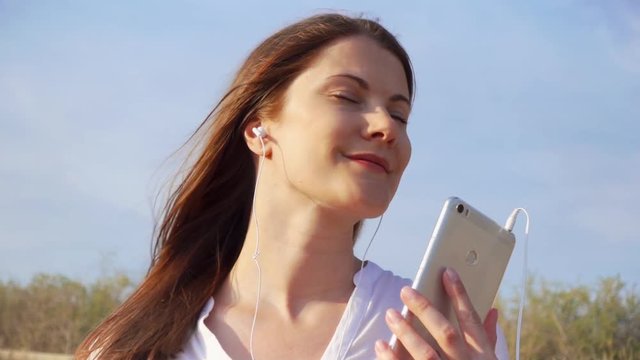 Young happy woman in white shirt using mobile against blue sky. Female listening music via app on cellphone. Wind blowing her hair in slow motion