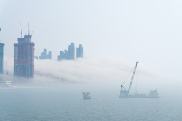 Thick large and heavy mist blow from the sea to the land and pass through shop near shore and skyscrapers inland and cover all other buildings in the area.