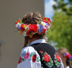 Dziewczyna (górna część ciała) stoi tyłem, włosy uczesane w warkocze, na głowie kolorowy kwietny wianek, ubrana w tradycyjny haftowany strój regionalny - obrazy, fototapety, plakaty