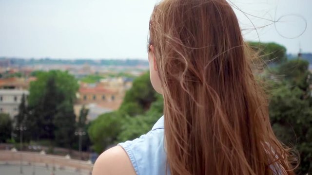 Young woman standing on observation deck looking at european city panorama. Carefree female traveler enjoying vacation in Europe alone. Girl's hair fluttering in wind in slow motion