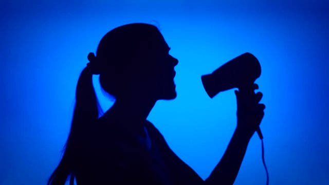 Silhouette of young woman having fun singing into hair dryer. Female's face in profile pretending to hear music dancing silly on blue background. Black contour shadow of teenager's half-face singing