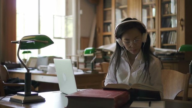Young female student lawyer is reading book, typing on laptop sitting in library, Asian cute woman in headphones is examining theme, using computer during learning week in vintage interior. Concept
