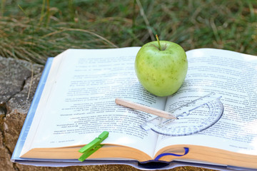 Fresh green apple on an open book outdoor in the grass. Time for school. Learn and healthy food concept. Apple and a book. Open schoolbook. Open manual and fruit outdoor. Nature and education. 