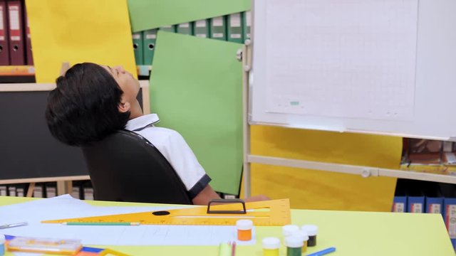 A boy sitting on a chair unwinds near a table with writing utensils