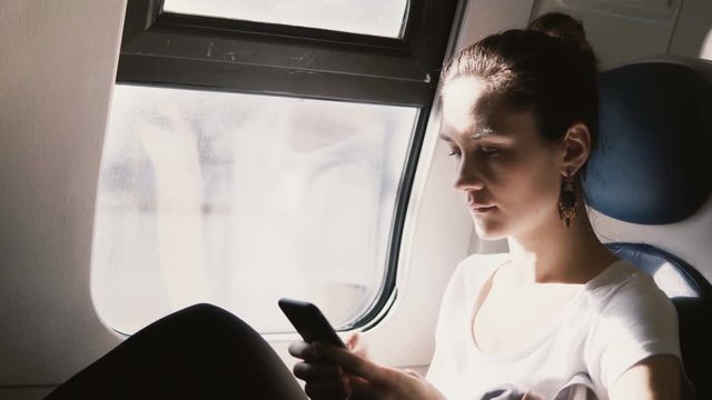 Beautiful peaceful European girl using smartphone app traveling in a train car, typing messages, looking at the window