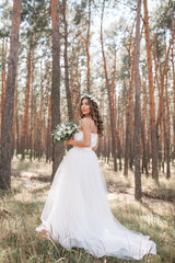 Obraz na płótnie Canvas A cute curly woman in a white wedding dress with a wedding bouquet and wreath in her hair standing back to the camera in nature. Concept escaped bride. Forward to a happy bright future Runaway