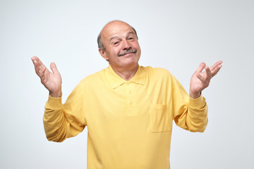 Indoor portrait of confused senior man in yellow t-shirt showing I have no idea gesture