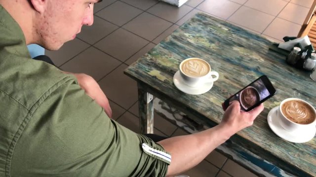 young athletic man, takes a picture of a cup with a coffee drink on a smartphone, resting sitting at a table in a cafe. communication in social networks, on the Internet