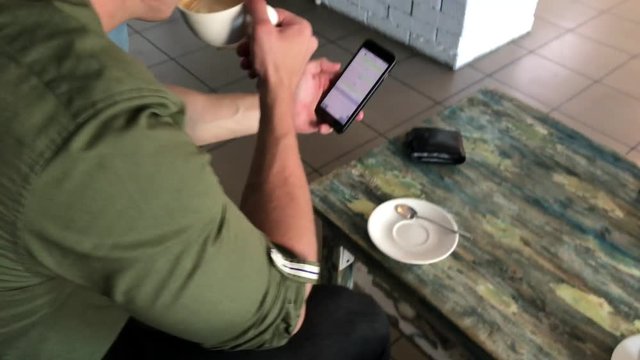 young athletic man, takes a picture of a cup with a coffee drink on a smartphone, resting sitting at a table in a cafe. communication in social networks, on the Internet