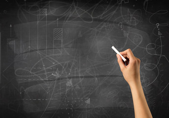 Female hand holding white chalk in front of a blackboard with scribbles and plans drawn on it 