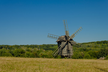 Fototapeta na wymiar Antique old wooden windmill under blue sky in front of forest. Sunny summer day. National Museum of Folk Architecture and Life of Ukraine in Pyrohiv. 