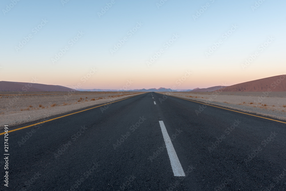 Wall mural Straight road though desert dunes towards sunset mountains, Sossusvlei, Namib  desert, Naukluft National Park, Namibia