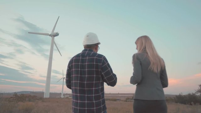 A young man in a helmet on the background of windmills talks about the project businesswoman. Work of windmills. The evening sky and the city are in the background.