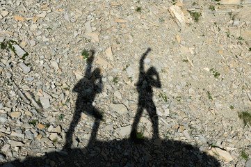 Two shadows of photographers on the earth going uphill with up-raised hands screaming about victory and success. Silhouettes of two people walking uphill