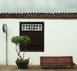 Tuinposter Beautiful detail of a typical facade of a small house with a big window a plant with flowers, tiles roof, a bench and a road sign in Garachico, Tenerife, Canary Island © vali_111