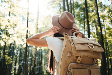 Young hipster woman traveling alone on outdoors among trees and hold hat his hands. Handsome girl...