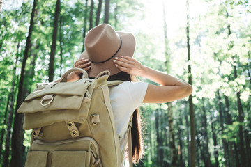 Back view on handsome traveler hipster girl with backpack and hat walking in forest among trees
