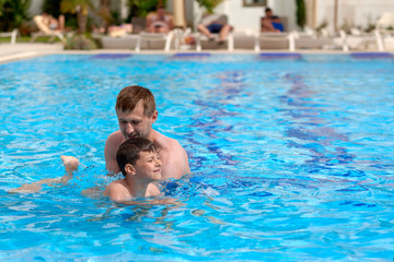 Naklejka premium Cute Caucasian boy is obtaining swimming skills in the swimming pool. His father is helping him.