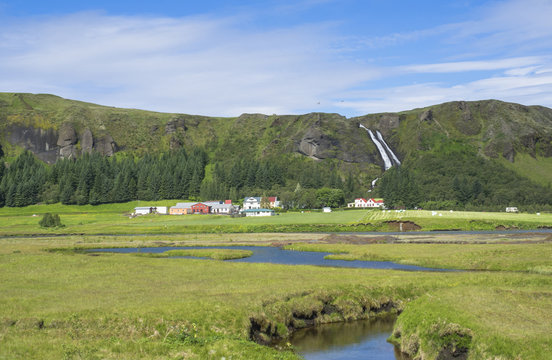 rural landscape with colorful farm houses, blue river stream on green grass meadow with spruce tree forest, hills and beatuful waterfall Systrafoss in Kirkjub&aelig;jarklaustur, Iceland