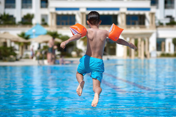 Smiling Caucasian boy in floating sleeves having fun in pool at resort.  HE is jumping into pool.