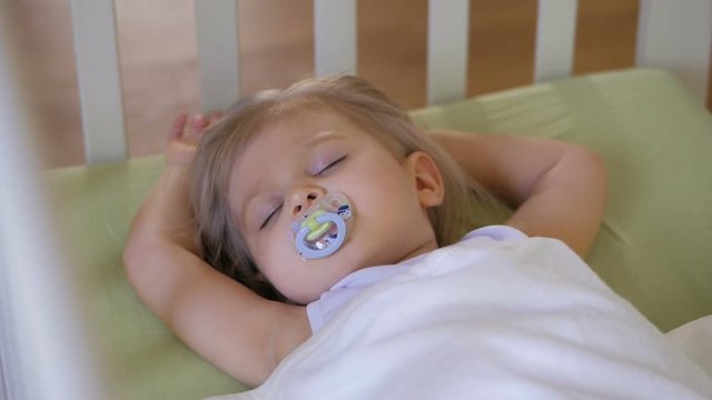 A little girl is sleeping in a baby crib with a pacifier in her mouth