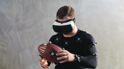 man holding american football rubber with motion sensors while using VR headset motion capture suit looking down throwing ball away virtual reality glasses sport skills developing in virtual reality - Powered by Adobe