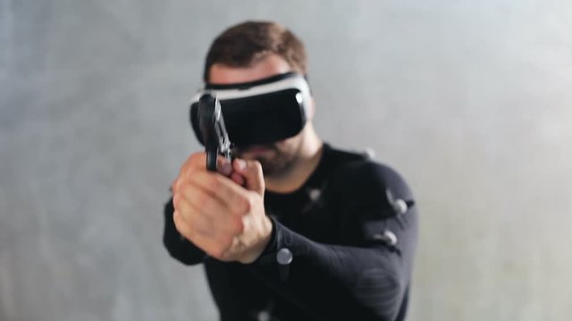 athletic bearded man playing VR shooter game with virtual reality headset testing motin capture suit while program recording the movement handgun pistol shooting closeup on grey background studio