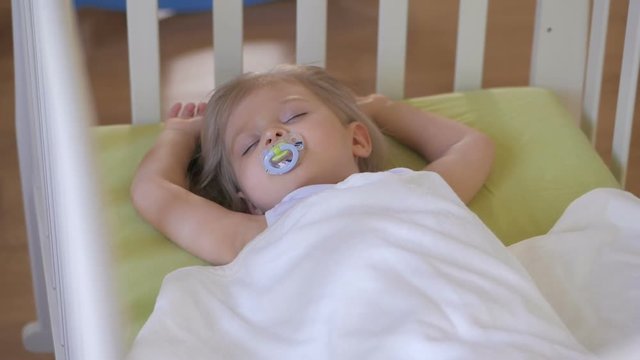 A little girl is sleeping in a baby crib with a pacifier in her mouth