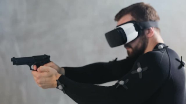 muscular adult man in virtual reality glasses and mocap suit with motion sensors shooting in cyberspace with real weapon gun holding 3d shooter interface design game develop industry male animator