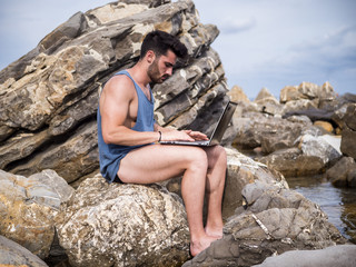 Young handsome man working on laptop computer, typing on keyboard while at the beach in front of the sea