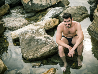 Handsome muscular young man sitting on a rocky beach, relaxed, shirtless, looking at camera