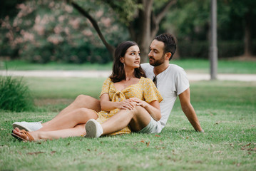 Fototapeta premium Stylish cute brunette girl sitting between legs of her boyfriend with beard close to each other on the grass in the park in Spain in the evening