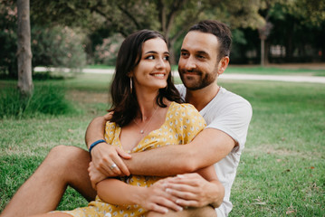 Naklejka premium Stylish young man with beard hugging his cute smiling girlfriend sitting between his legs on the grass in the park in Spain in the evening