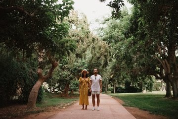 Stylish cute brunette girl with her boyfriend with beard posing holding hands close to each other on the small road in the park between large trees in Spain in the evening. Date of the young couple