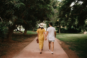 Stylish cute brunette girl with her boyfriend with beard walking holding hands close to each other on the small road in the park between large trees in Spain in the evening. Date of the young couple