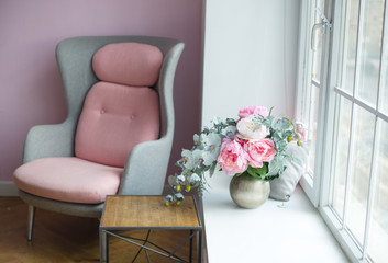 gray chair with pink insert stands near the window, on the window vase with pink peonies